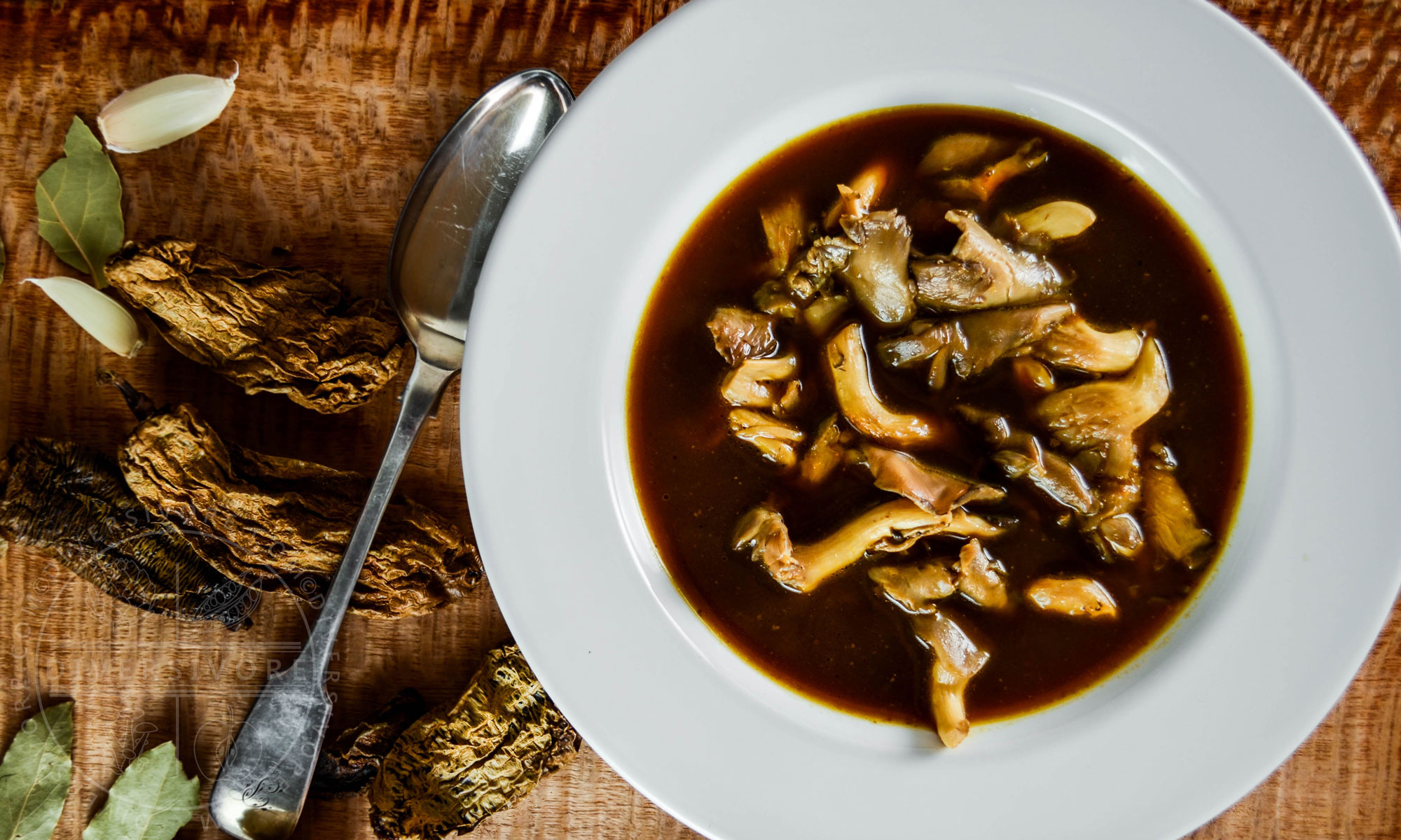 Smoky Vegan Mexican Oyster Mushroom Soup beside a spoon, bay leaves, garlic, and chipotle meco peppers.