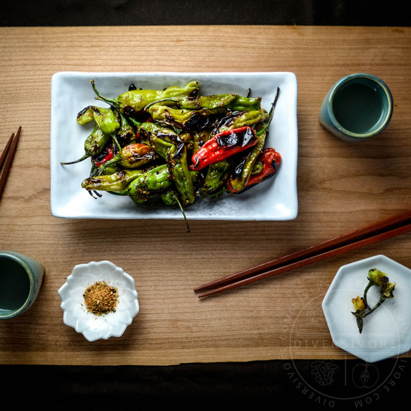 Charred shishito peppers with a sweet and spicy shichimi togarashi glaze on a white plate with chopsticks, tea cups, and small side plates