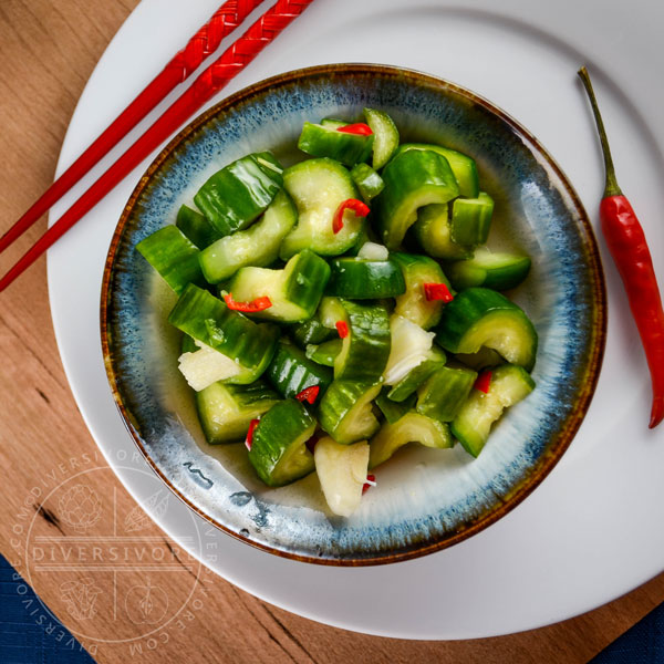 Chinese garlic cucumbers (Liangban Huanggua) in a small ceramic bowl, on a plate with red chopsticks and a chili pepper