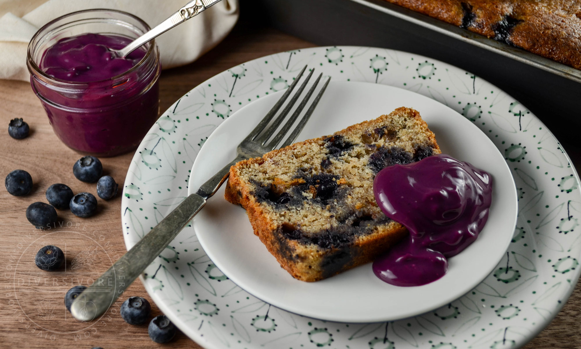 A slice of blueberry juniper rye cake on a white plate with an old fork, topped with blueberry-lemon curd