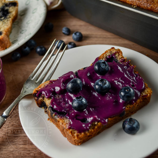 A slice of blueberry juniper rye cake on a white plate with an old fork, topped with blueberry-lemon curd and fresh blueberries
