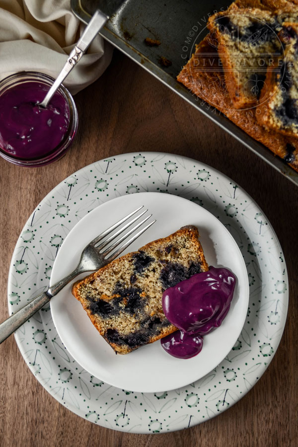 A slice of blueberry juniper rye cake on a white plate with an old fork, topped with blueberry-lemon curd