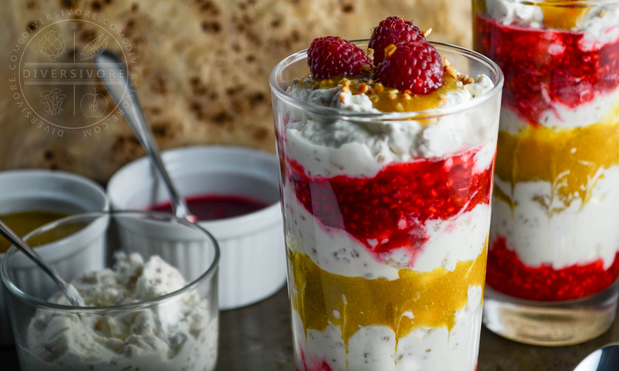 Cranachan with raspberries and greengages in two tall glasses, served in front of bowls of extra sauce and whipped cream