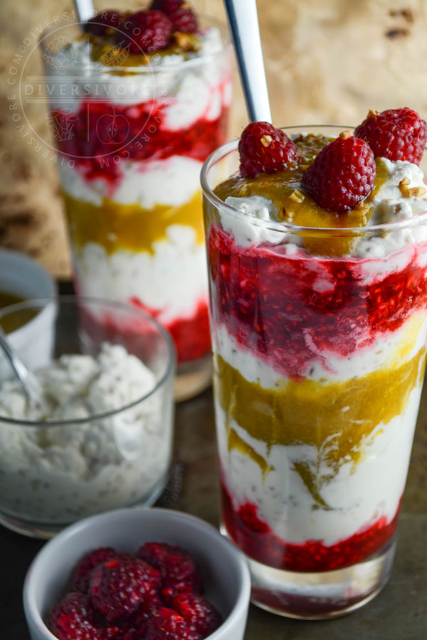 Cranachan with raspberries and greengages in two tall glasses, served in front of bowls of extra sauce and whipped cream