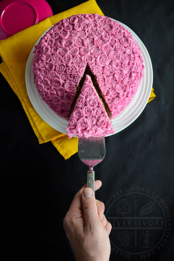 A hand holding a cake lifter removes a slice from a chocolate cake made with beetroot and candied pecans, frosted with pink cream cheese frosting rosettes