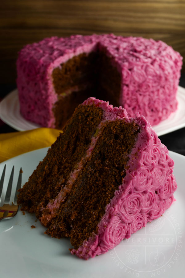 A slice of chocolate cake made with beetroot and candied pecans, frosted with pink cream cheese frosting rosettes, shown in front of the whole cake.