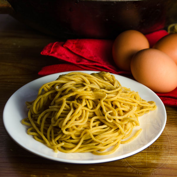 Homemade Chinese egg noodles on a white plate, in front of a red cloth and brown eggs.