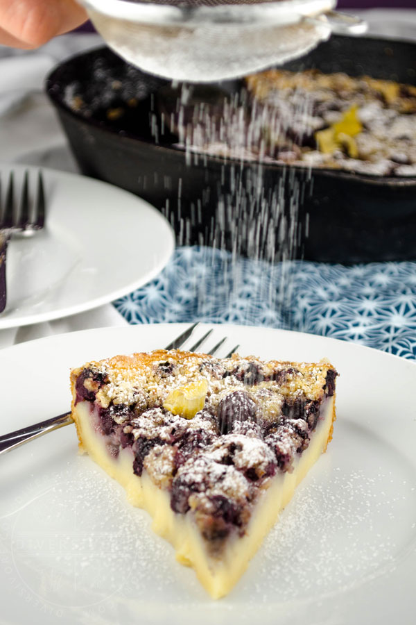 Saskatoon berry clafoutis topped with candied lemon peel being dusted with powdered sugar, on a white plate with a fork