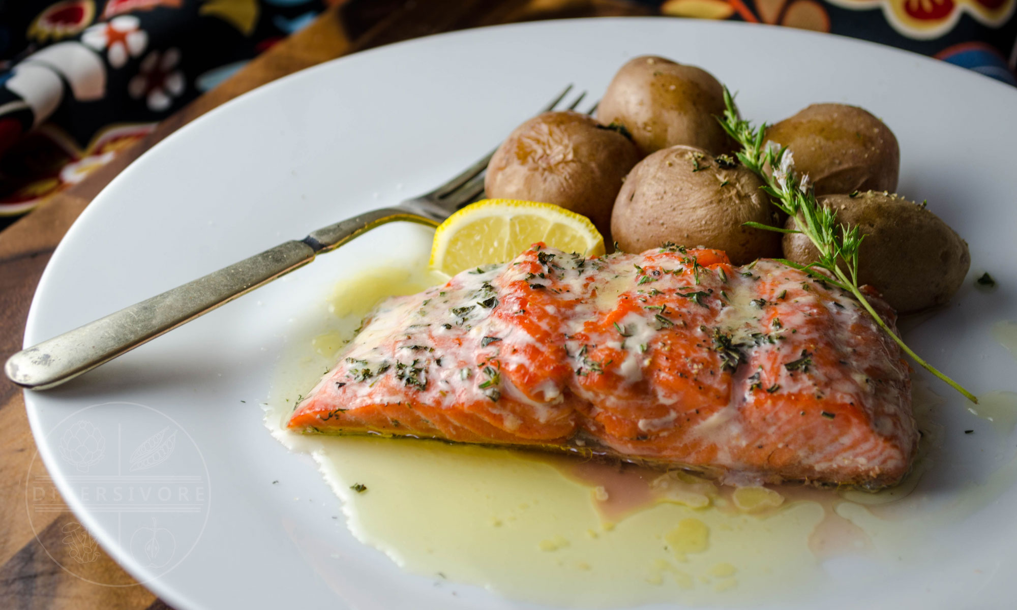 Salmon with fresh herbs and lemon-garlic butter, served on a white plate with new potatoes and a sprig of winter savoury