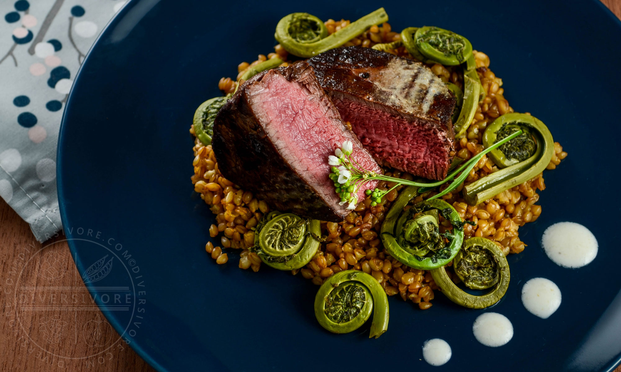 Seared beef tenderloin medallions with simmered einkorn (heirloom wheatberries), sauteed fiddleheads, and a horseradish-yogurt cream on a dark blue plate