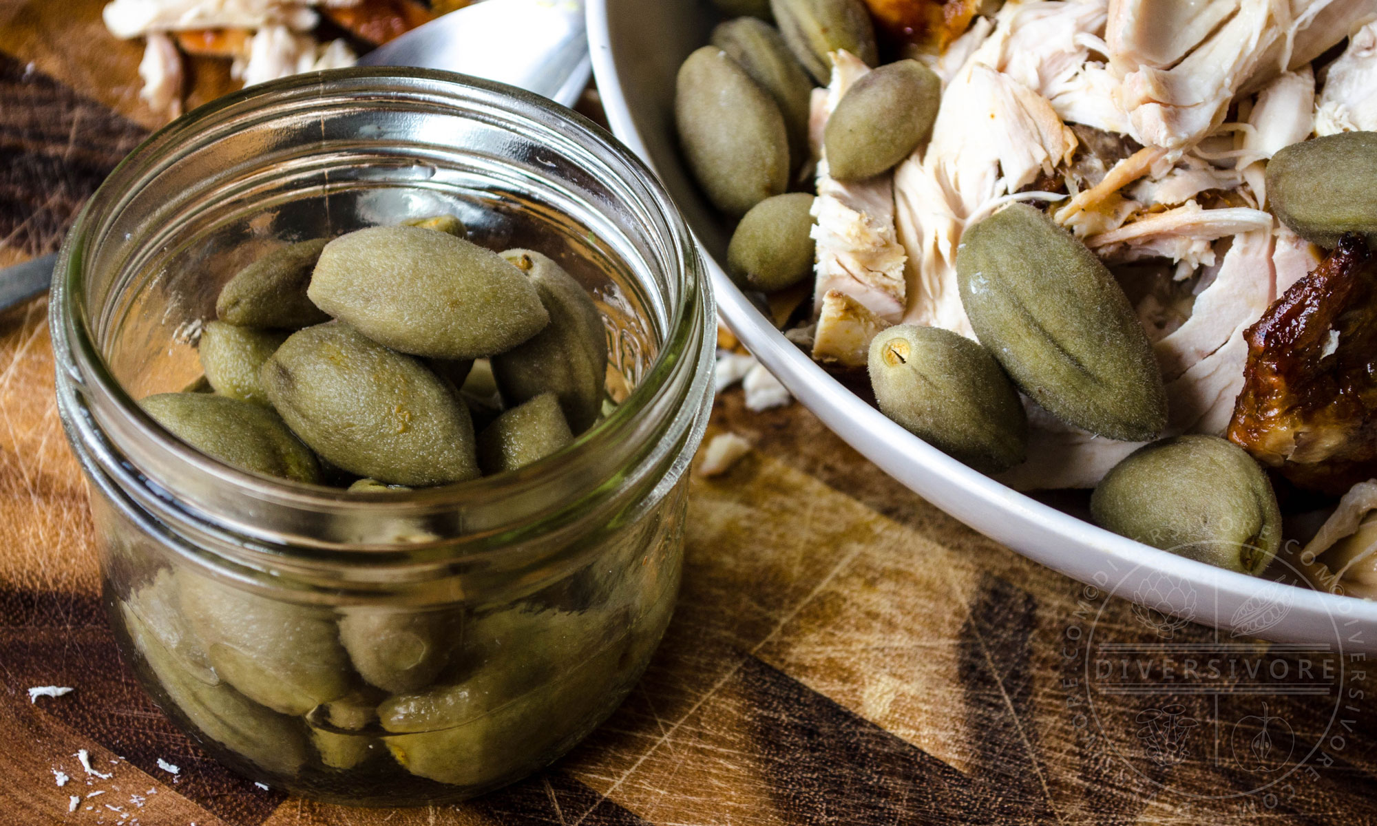 Pickled green almonds in a glass mason jar in front of a plate of chicken