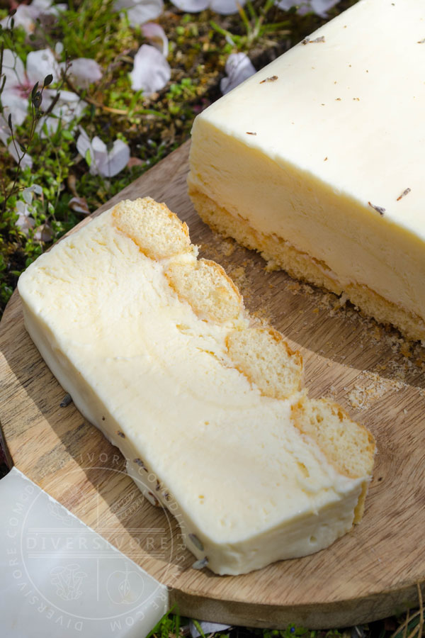 Easy Lemon and Lavender Semifreddo with one slice cut off, shown on a wood board against a backdrop of cherry petals and grass
