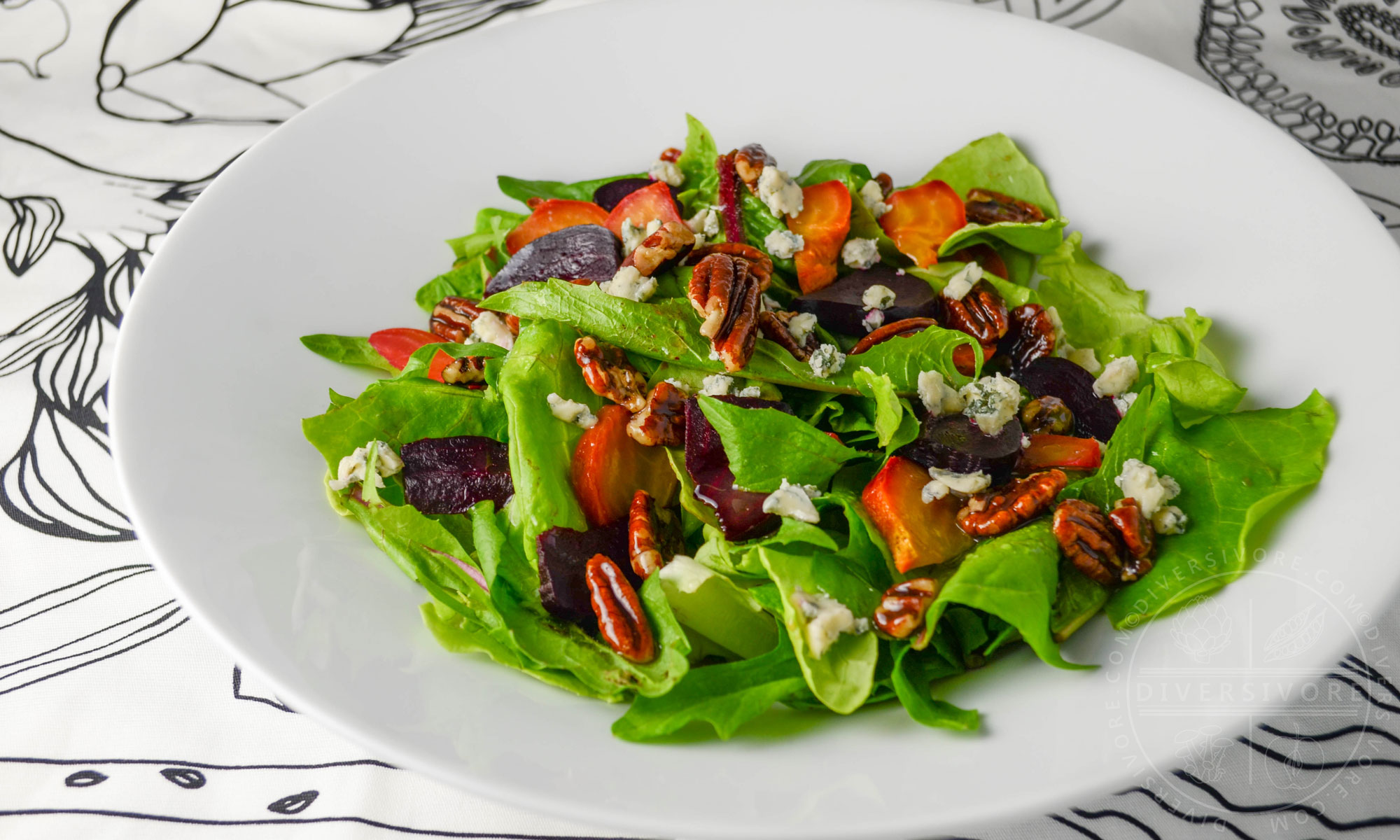 Dandelion and butterleaf lettuce salad with roasted beets, blue cheese, and maple-candied pecans in a white bowl against a black and white patterned cloth