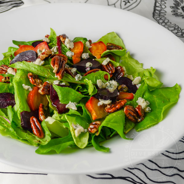 Dandelion and butterleaf lettuce salad with roasted beets, blue cheese, and maple-candied pecans in a white bowl against a black and white patterned cloth