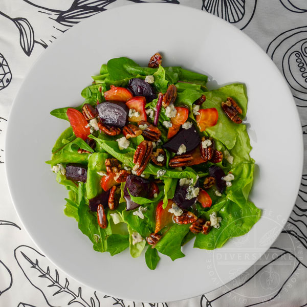 Dandelion and butterleaf lettuce salad with roasted beets, blue cheese, and maple-candied pecans in a white bowl against a black and white patterned cloth