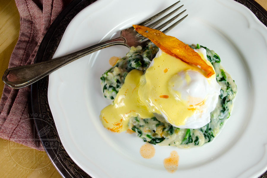 Eggs Sardou (poached egg with Hollandaise on an artichoke bottom with creamed spinach and artichokes), topped with a fried tomato skin and served on a white plate with a silver fork