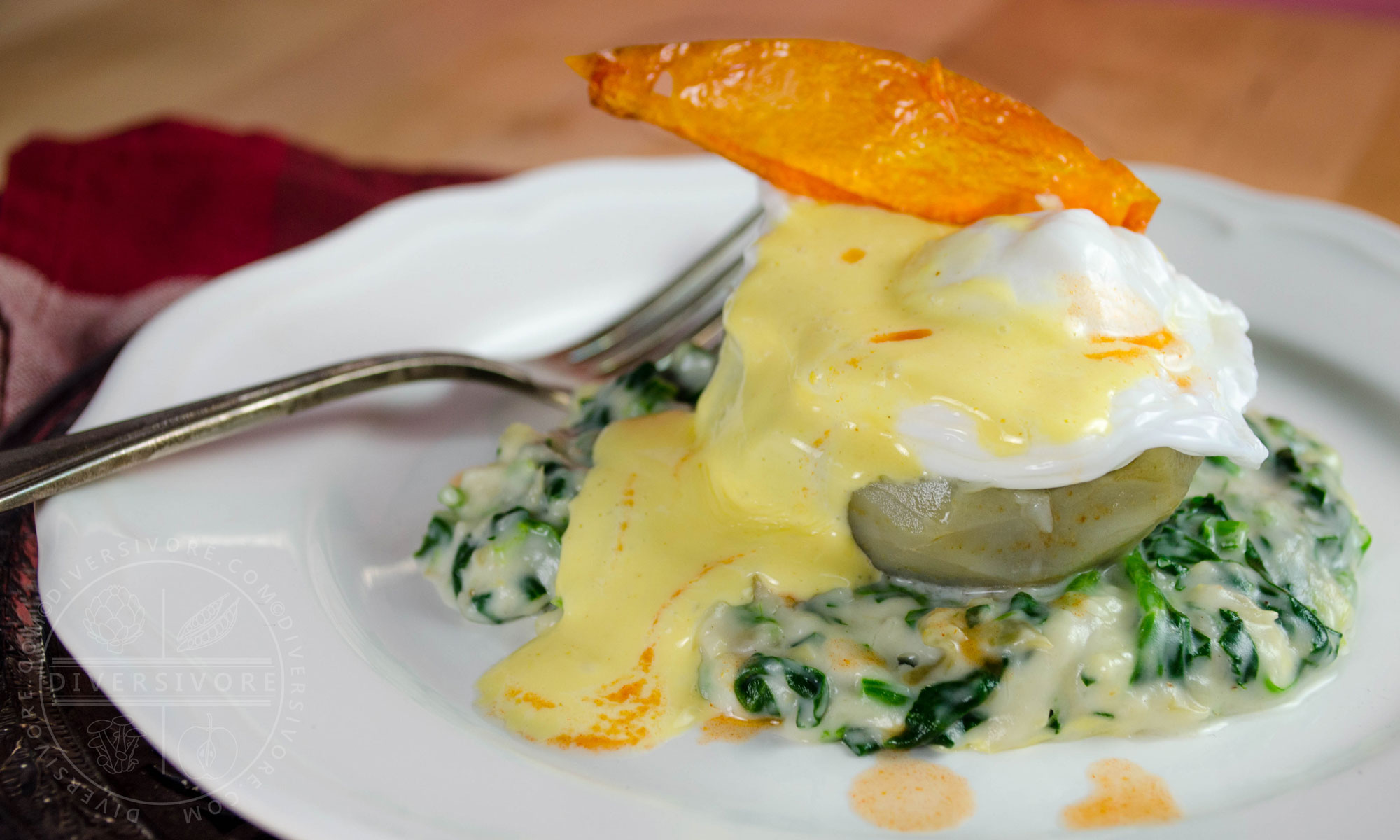 Eggs Sardou (poached egg with Hollandaise on an artichoke bottom with creamed spinach and artichokes), topped with a fried tomato skin and served on a white plate with a silver fork