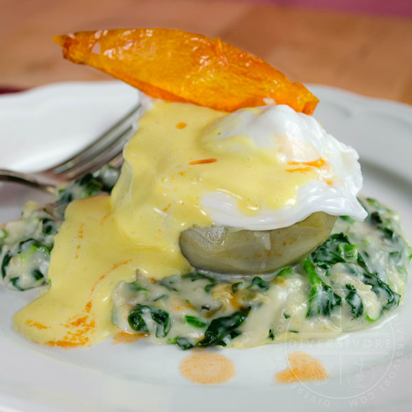 Eggs Sardou (poached egg with Hollandaise on an artichoke bottom with creamed spinach and artichokes), topped with a fried tomato skin and served on a white plate with a silver fork