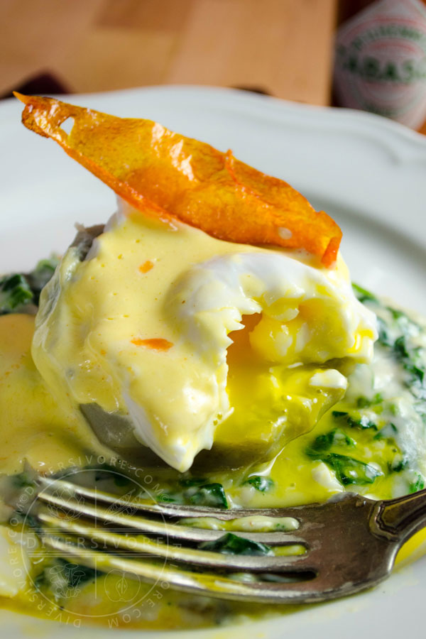 Eggs Sardou (poached egg with Hollandaise on an artichoke bottom with creamed spinach and artichokes), topped with a fried tomato skin and served on a white plate with a silver fork