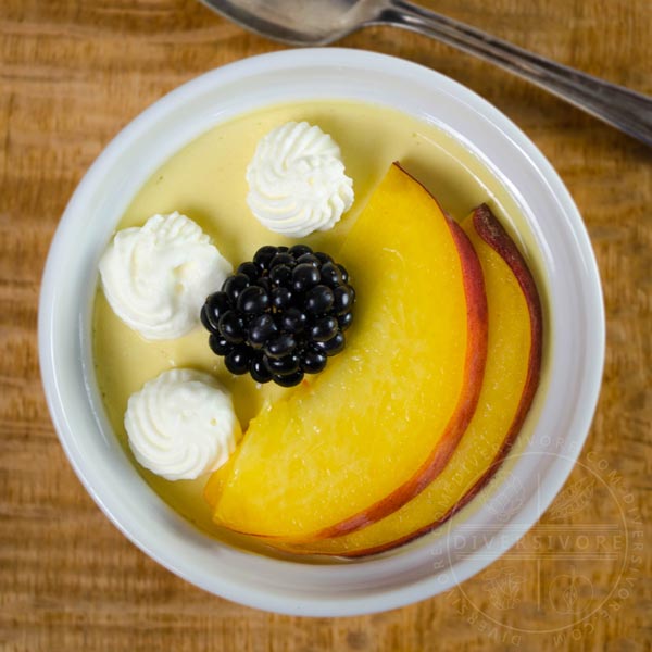 An overhead view of a ramekin of peach pot de creme with peach slices, blackberry, and whipped cream on a wood background