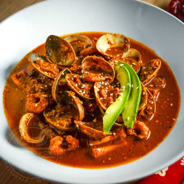 Red Pipian seafood soup with avocado slices in a white bowl