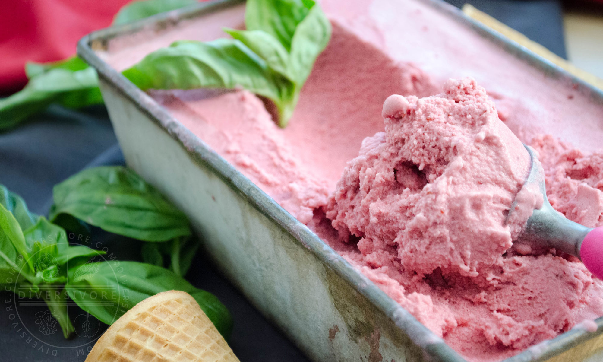 A scoop being taken out of a container of strawberry, basil, and goat cheese ice cream, decorated with basil leaves