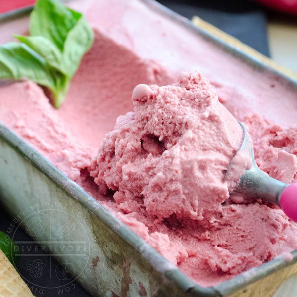 A scoop being taken out of a container of strawberry, basil, and goat cheese ice cream, decorated with basil leaves