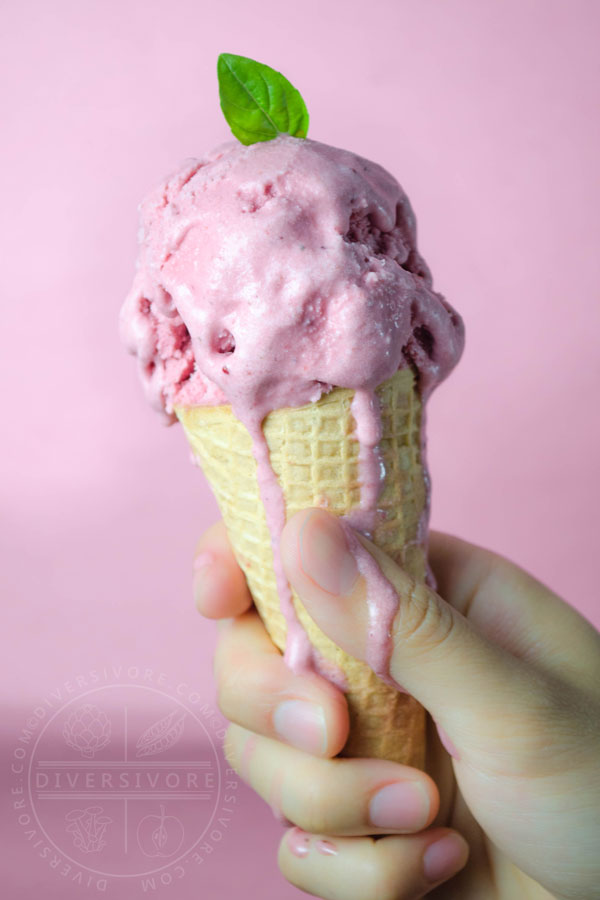 Strawberry, basil, and goat cheese ice cream dripping down an ice cream cone and onto a hand against a pink backdrop