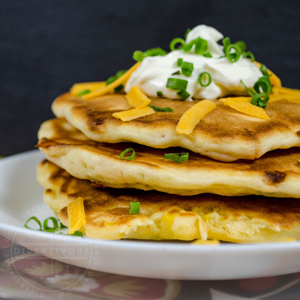 A stack of bacon, cheddar, and chive pancakes topped with sour cream and cheese on a white plate