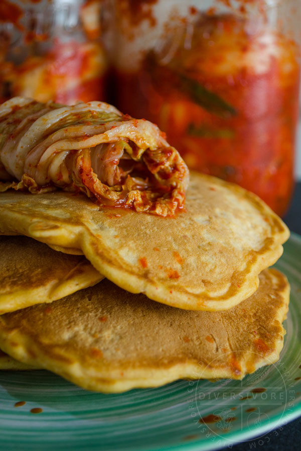 Kimchi and seafood pancakes on a green plate, topped with kimchi, in front of kimchi jars