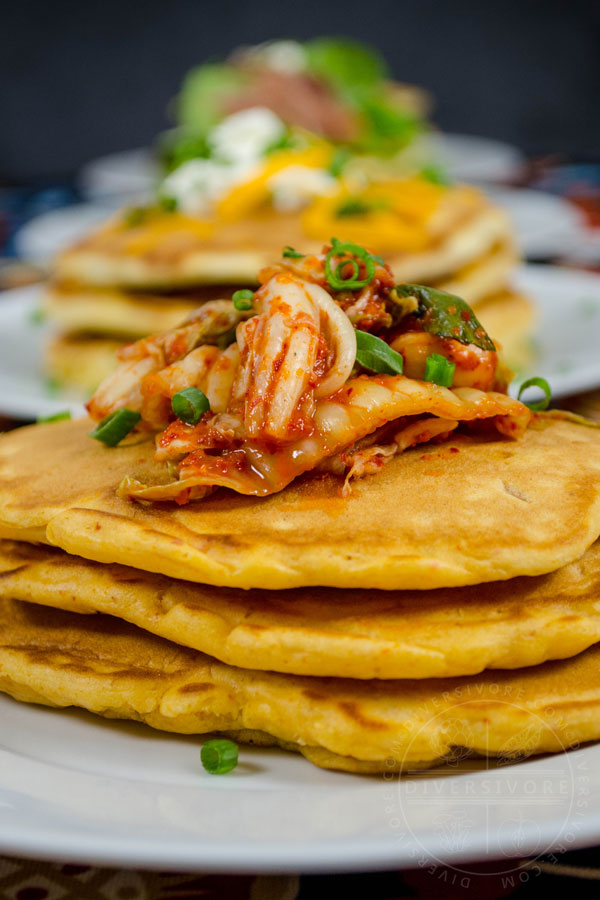Kimchi and seafood pancakes on a green plate, topped with kimchi and green onions, served in front of other savory pancake variations.