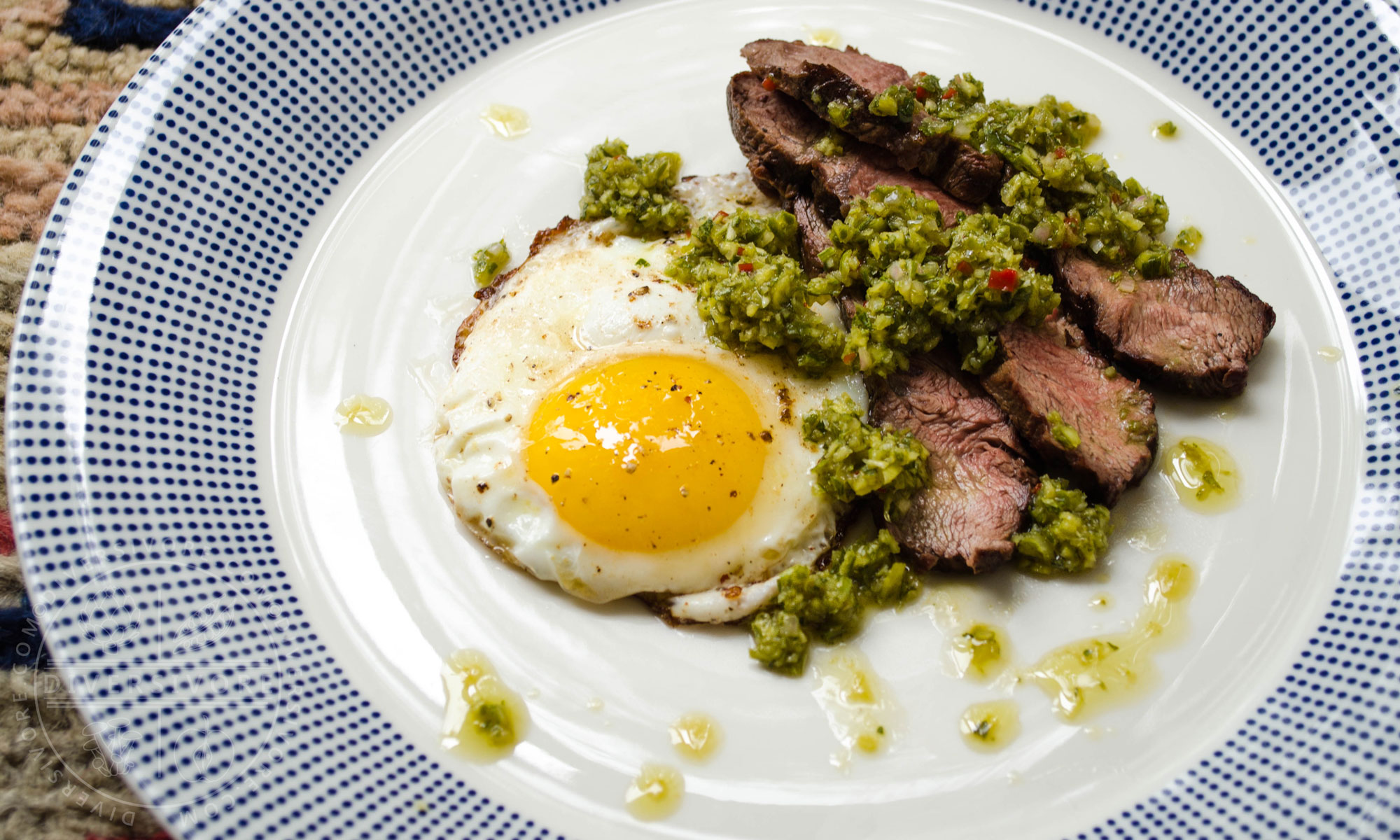 Garlic scape chimichurri on top of sliced steak and a fried egg, served on a white and blue patterned plate