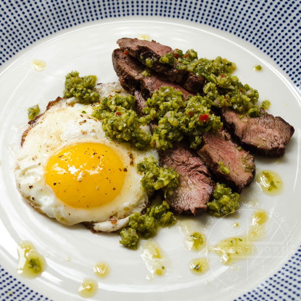 Garlic scape chimichurri on top of sliced steak and a fried egg, served on a white and blue patterned plate