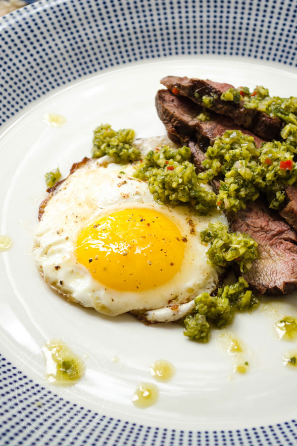 Garlic scape chimichurri on top of sliced steak and a fried egg, served on a white and blue patterned plate