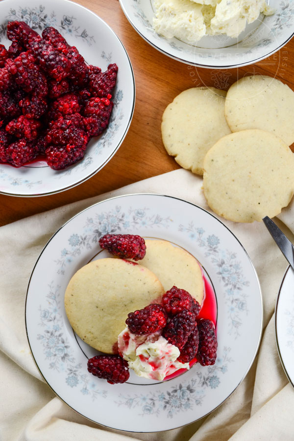 Pepper shortbread cookies with tayberries and sweet cream cheese on a small floral patterned plate, flanked by additional cookies and a bowl of tayberries