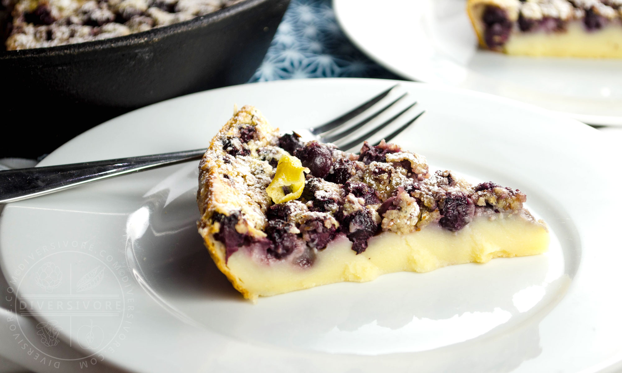 Saskatoon berry clafoutis topped with candied lemon peel and powdered sugar, on a white plate with a fork