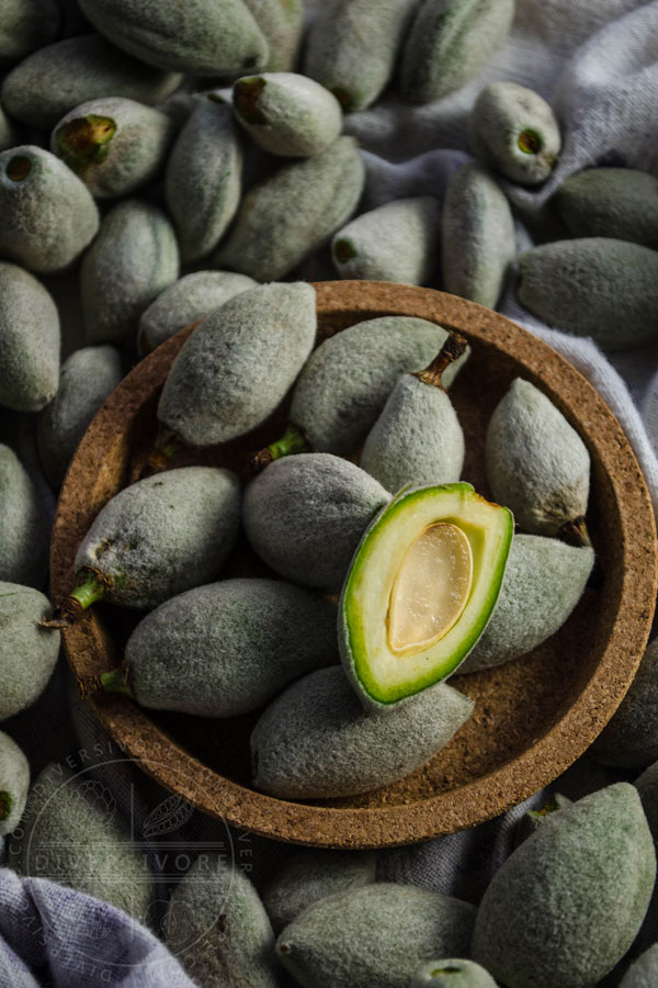 Green almonds, including one cut in half, in and around a small cork bowl