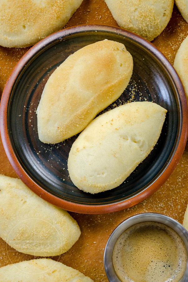 Two pandesal rolls on a plate, surrounded by more bread, with a small coffee cup