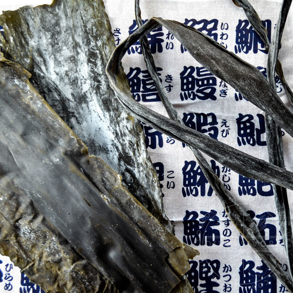 Different types of kombu (dashi kelp) in front of a cloth covered with Japanese writing.
