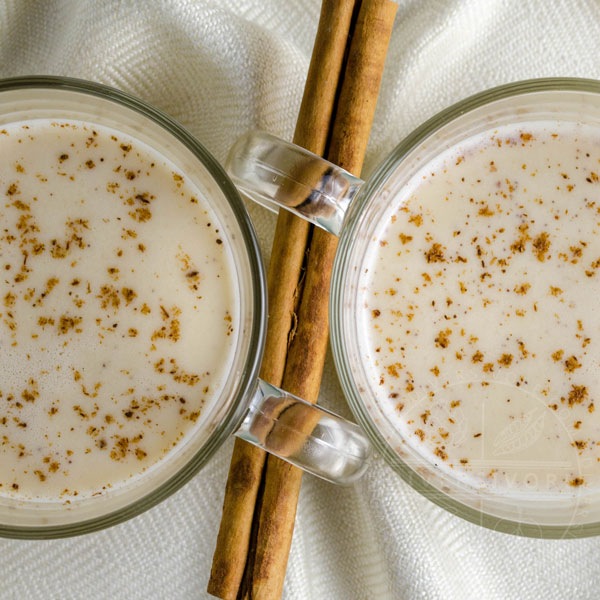 Two mugs of coquito, dusted with cinnamon, and separated by a cinnamon stick